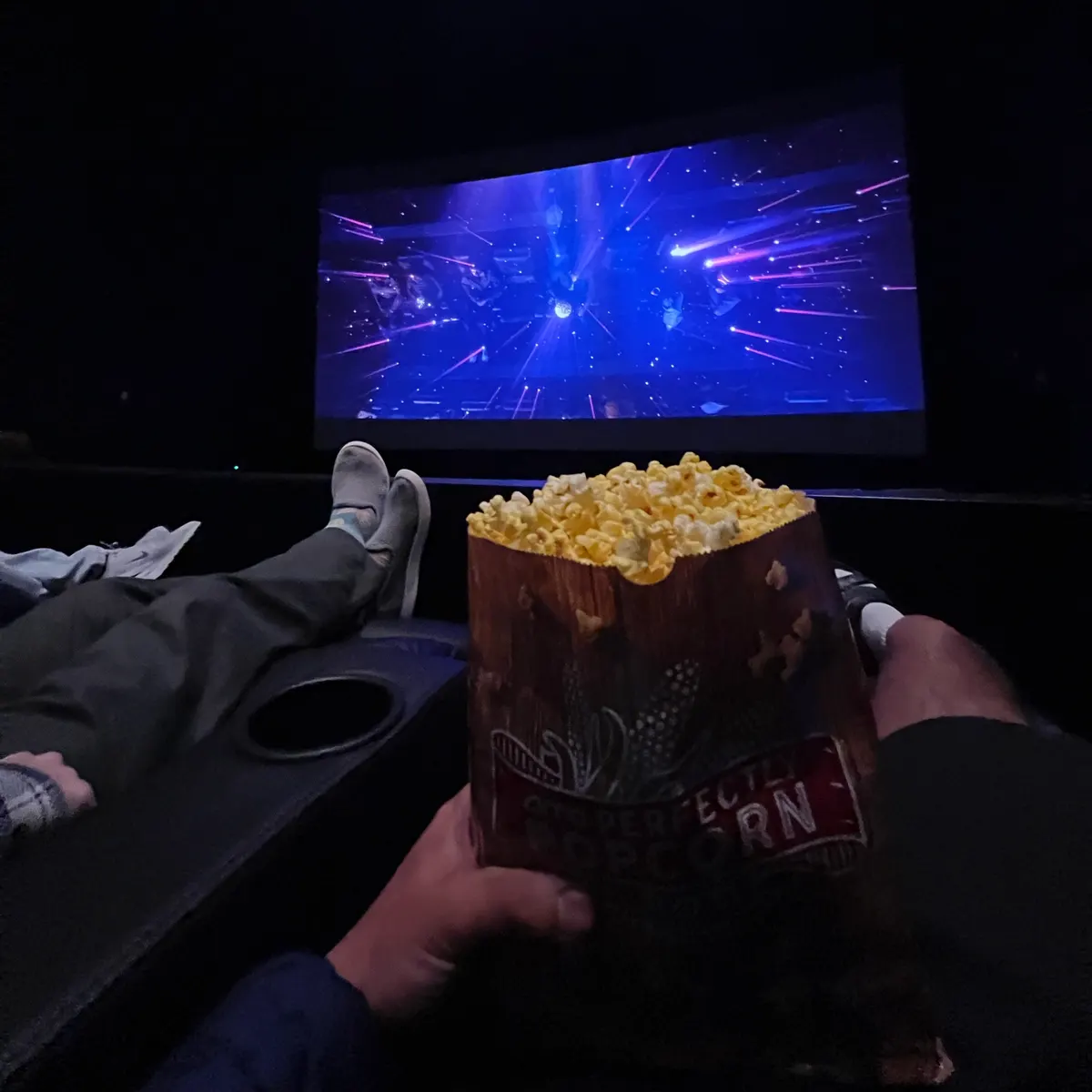 A bag of popcorn held up in a dark cinema, feet up, with the screen glowing blue