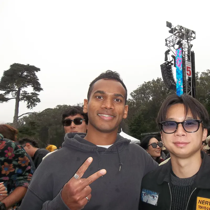 Two friends smiling at an outdoor festival, one making a peace sign
