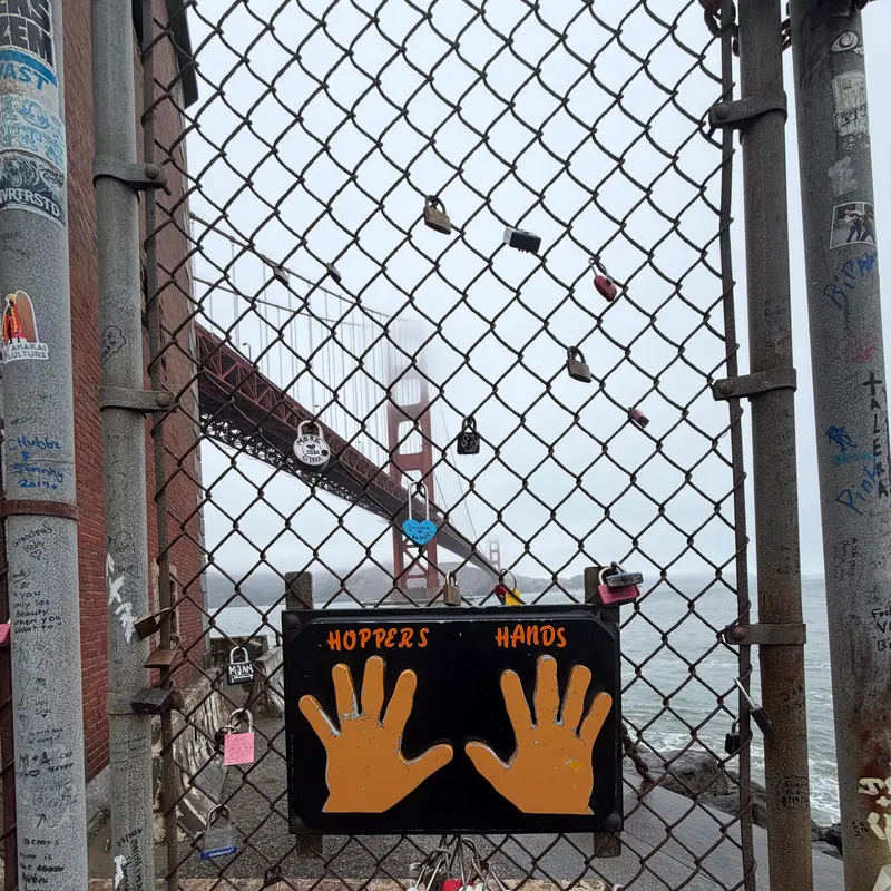 The Golden Gate Bridge in fog, seen through a chain-link fence with a Hopper’s Hands sign