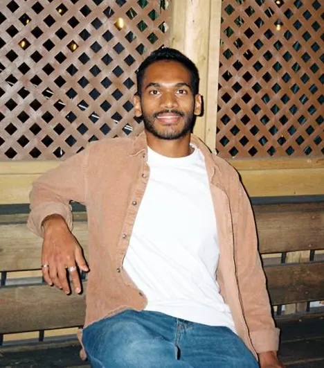 Sathira Katugaha smiling, sitting on a wooden bench in a tan corduroy shirt and white T-shirt